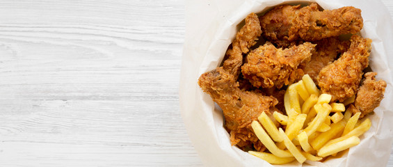 Fast food: fried chicken drumsticks, spicy wings, French fries and chicken strips in paper box over white wooden surface, top view. Copy space.