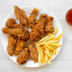 Tasty fried chicken drumsticks, spicy wings, French fries, chicken tenders and sauce on white plate over white wooden surface, top view. Flat lay, overhead, from above.