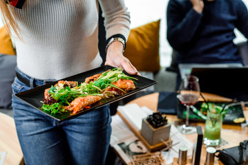 Woman in jeans and a gray sweater in a restaurant is holding a black plate with vegetable spring rolls. Close up. Space