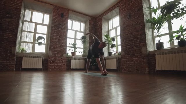 Young man doing an asana in slow motion. Yogi master workout on black mat on urban studio. Guy doing yoga indoors near windows. Healthy lifestyle concept. Slider, dolly shot with copy space