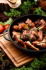 Roasted chicken legs in a black cauldron on a wooden board among fresh vegetables on a dark wooden background. Vegetable composition. Chicken BBQ. Close-up.