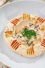 Hummus with greens in a round white plate on a light wooden background among fresh vegetables. Close-up. Space. Middle Eastern cuisine. 