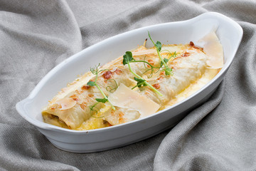 Cannelloni with minced meat, baked in a white ceramic plate. On textile background