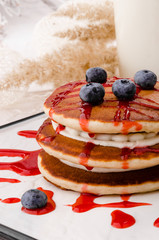 Beautiful pancakes with blueberries and red jam on a white square plate and milk on a light spring background, decorated with fresh roses. Concept of a colorful and healthy breakfast. Close-up. Space