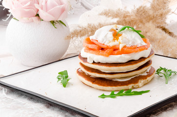 Beautiful pancakes with salmon, poached egg and arugula on a white plate on a light background, decorated with pink roses. Close-up. Space