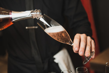 Waiter in black shirt is pouring rose champagne in wine glass. Closeup on hands and glass.