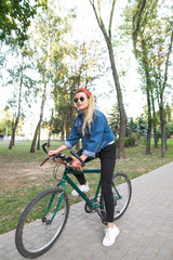 Attractive girl in stylish clothes sitting on a bike in the park. Teenage girl in sunglasses and a denim jacket is in a park with a bicycle. Walk on the bike.