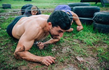 Male participant in an obstacle course crawling under electrified cables with partners in background