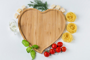 on a white background a wooden plate in the shape of a heart, around it are tomatoes, olive oil, dill, cheese, basil and pasta in the form of nests 
