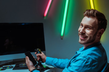 selective focus of smiling and handsome man holding joystick and smartphone