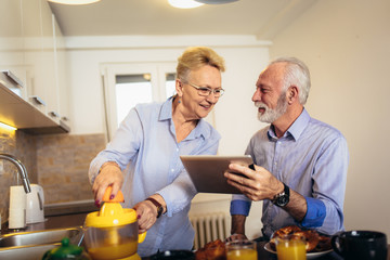 Aged couple busy look at digital tablet while having delicious breakfast at home kitchen