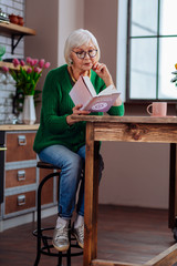 Aging woman being absorbed in book sitting at furnished kitchen