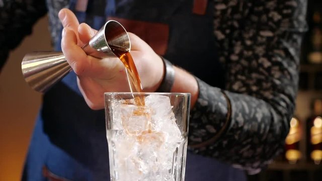 Bartender hands measuring cocktail ingredient in special cup or jigger then pouring it in a glass over ice - close up, slow motion