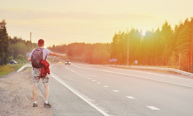 Hitchhiking young traveller man try to catch car on a forest road in sunset. Caucasian tourist guy...