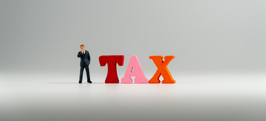 miniature figure businessman in dark blue suit standing beside colorful wooden TAX word and thinking abount TAX.