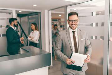 Young Caucasian businessman in formal wear and with eyeglasses posing with clipboard in his hand...