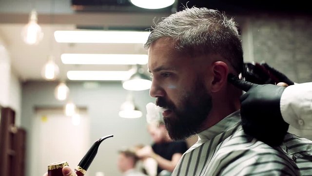 Hipster man client visiting haidresser and hairstylist in barber shop, smoking a pipe.