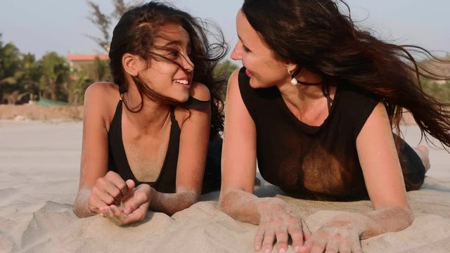 Portrait of happy mother and teen daughter lying together on the sandy beach, talking and laughing in slow motion.