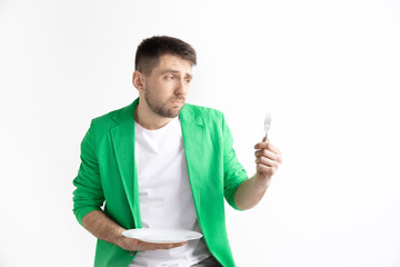 Young sad attractive caucasian guy holding empty dish and fork isolated on grey background. Copy space and mock up. Blank template background. Reject, rejection concept