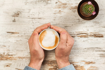 Manos de hombre sosteniendo una taza de café con leche sobre una mesa de madera blanca rústica....