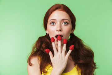 Beautiful emotional young redhead girl posing isolated over green wall background with raspberry.