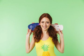 Happy young redhead girl posing isolated over green wall background with skateboard.