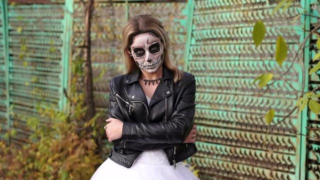 A young girl with a creepy make-up in the form of a skull on her face in a wedding dress and a leather jacket on the background of an old rusty fence. Halloween. The image of the dead bride.