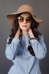 Sensual brunette model with round sunglasses, curly hair, wearing in jeans shirt and beige hat, touches his neck.