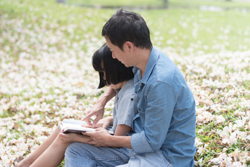 Asia male parent dad read a booking with his girl in garden. Asian father reading a story with his daughter in green public outdoor park. Family concept. National Father's day