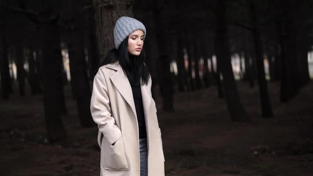 attractive girl in knitted hat and long spring coat walks through old pine wood in morning slow motion