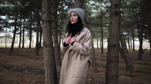 pretty elegant lady in knitted hat and light coat stands near young pine trees in spring wood slow motion