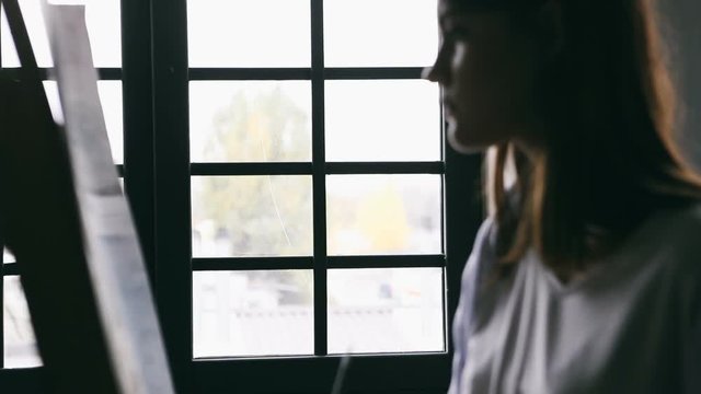 Painter girl is drawinf in front of the window. White short. Creative mood moment. Video footage