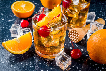 Classic alcohol cocktail, whiskey with ice cubes, orange slices and cocktail red cherries. In two glasses, on a dark blue concrete table,