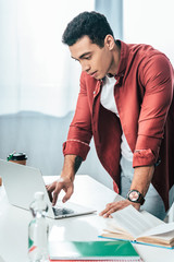 Concentrated brunette student in red shirt using laptop at workplace