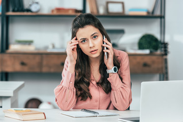 Serious attractive girl in pink shirt sitting at table and talking on smartphone