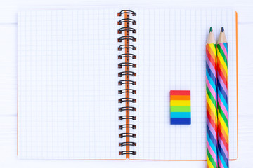 Open copybook with two rainbow-coloured pencils and a rubber on white wooden table