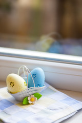 Colorful blue and yellow Easter eggs in basket with decorative flower near window in daylight.