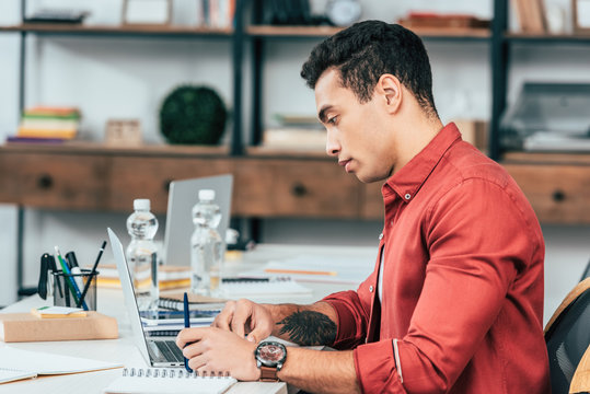 Concentrated Student With Tattoo In Red Shirt Using Laptop At Desk