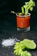 Cocktail Bloody Mary with lime and celery, salt and snacks in a glass on a black wooden table. vertical view of a cocktail.