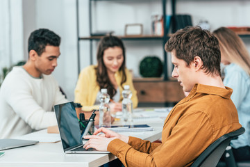 Student sitting at table with multiethnic friends and using laptop