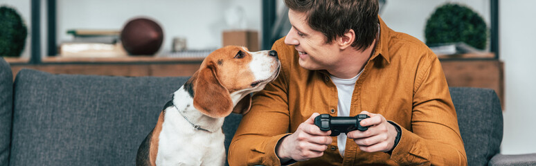 Panoramic shot of smiling young man with gamepad sitting on sofa and looking at dog
