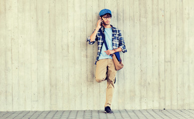 leisure, technology, communication and people concept - smiling hipster man with shoulder bag calling on smartphone at street wall