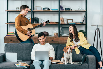 Three multicultural students with dog using laptops and playing guitar in living room
