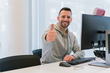 Young man with thumbs up happy and smiling for having achieved his goals in the office in front of the computer