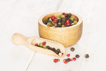 Lot of whole peppercorns of four colors with wooden bowl and wooden scoop on white wood