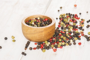 Lot of whole peppercorns of four colors with wooden bowl on white wood