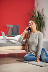 Obraz premium Beautiful Caucasian brunette dressed casual sitting on floor and leaning on bed in bedroom while talking on the phone. On bed laptop.