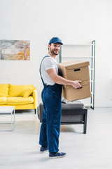 mover in uniform looking at camera while carrying cardboard boxes in apartment