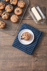 Homemade Blueberry Muffins on Wooden Table Top; One Isolated on White Plate on Blue Kitchen Towel; More Muffins on Cooling Rack in Background; Butter Dish and Knife beside the Muffin 