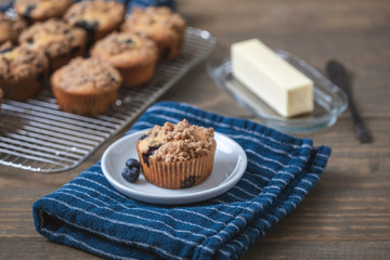 Homemade Blueberry Muffins on Wooden Table Top; One Isolated on White Plate on Blue Kitchen Towel; More Muffins on Cooling Rack in Background; Butter Dish and Knife beside the Muffin 